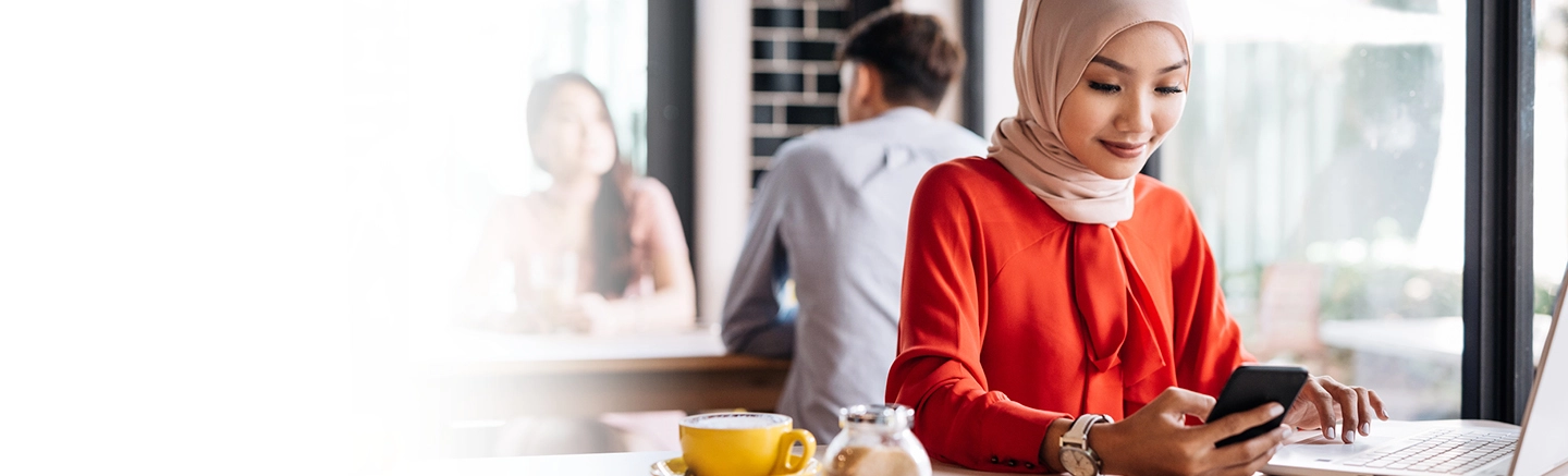 Personne portant un haut rouge à manches longues et un foulard clair, assise à une table de café avec un ordinateur portable et un téléphone intelligent. Une tasse de café jaune et un petit pot sont sur la table, et l’arrière-plan montre d’autres personnes et de grandes fenêtres.