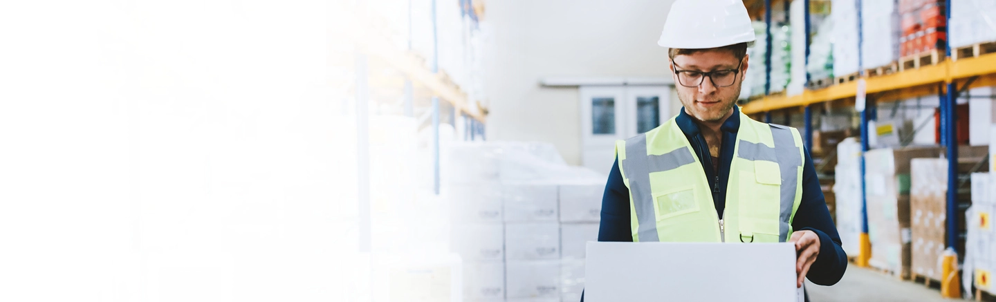 Warehouse worker wearing a white safety helmet and high-visibility vest, holding a large box in a storage area with shelves stocked with various packages and products.