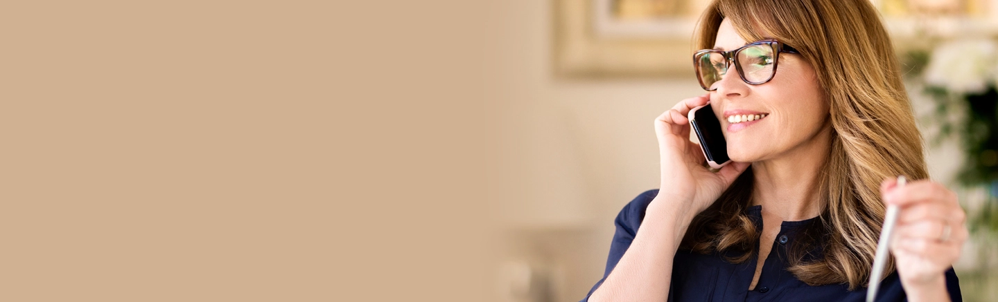 Person holding a smartphone to the ear while stirring a drink with a straw, seated indoors in a softly lit setting with a neutral background.