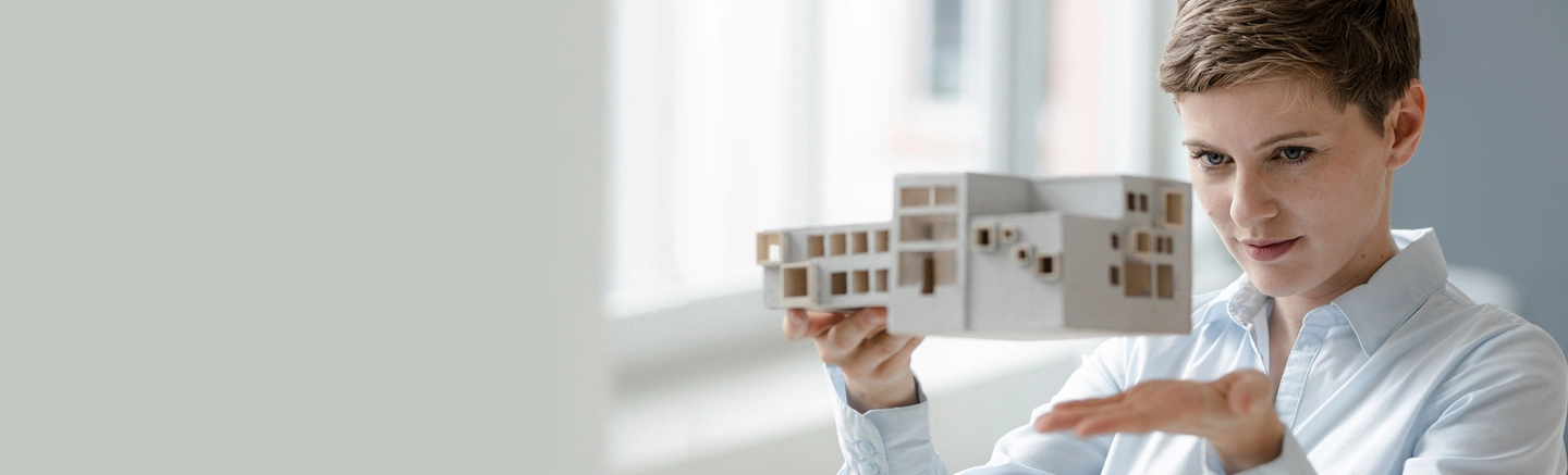 Personne tenant une maquette architecturale détaillée d’un bâtiment moderne, assise à l’intérieur près de grandes fenêtres laissant entrer la lumière naturelle.