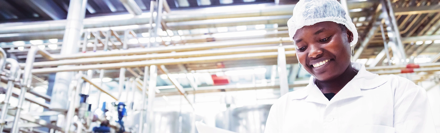 Travailleur portant une blouse blanche et un filet à cheveux dans une installation industrielle moderne avec des cuves en acier inoxydable, des tuyaux et des équipements utilisés pour la transformation alimentaire ou chimique.