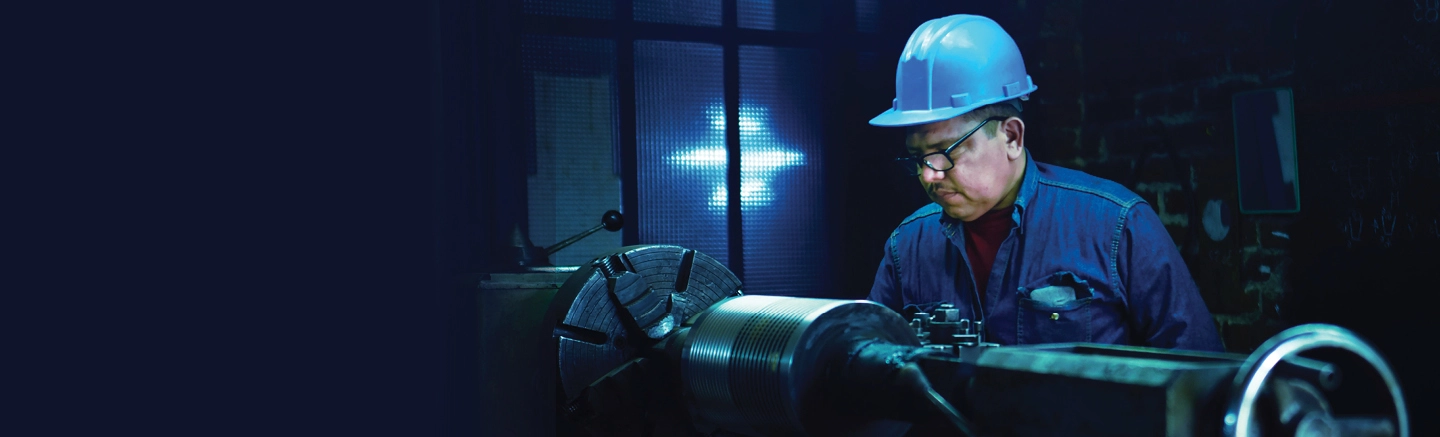 Industrial worker wearing a blue safety helmet and protective clothing, operating heavy machinery in a dimly lit workshop with metal equipment and a large rotating component.