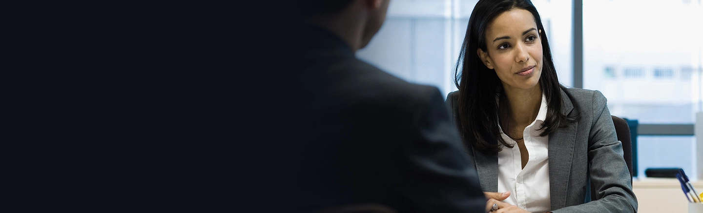 Deux personnes assises à un bureau dans un environnement professionnel, en pleine conversation. L’une porte un costume d’affaires, et l’arrière-plan comprend de grandes fenêtres laissant entrer la lumière naturelle.