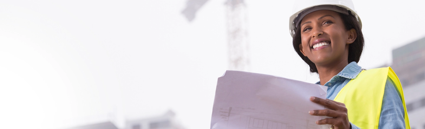 Jobseeker Construction HeadeTravailleur de la construction portant un casque de sécurité bleu et un gilet réfléchissant, debout dans un grand intérieur de bâtiment non terminé et tenant des plans architecturaux. L’espace présente des planchers en béton, des colonnes blanches et des tuyaux apparents au plafond.r