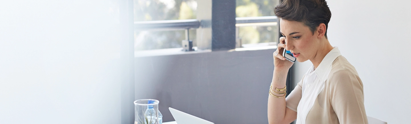 Person sitting at a desk in a bright office space, holding a smartphone near the ear, with a laptop and a glass of water on the table. Large windows in the background provide natural light.