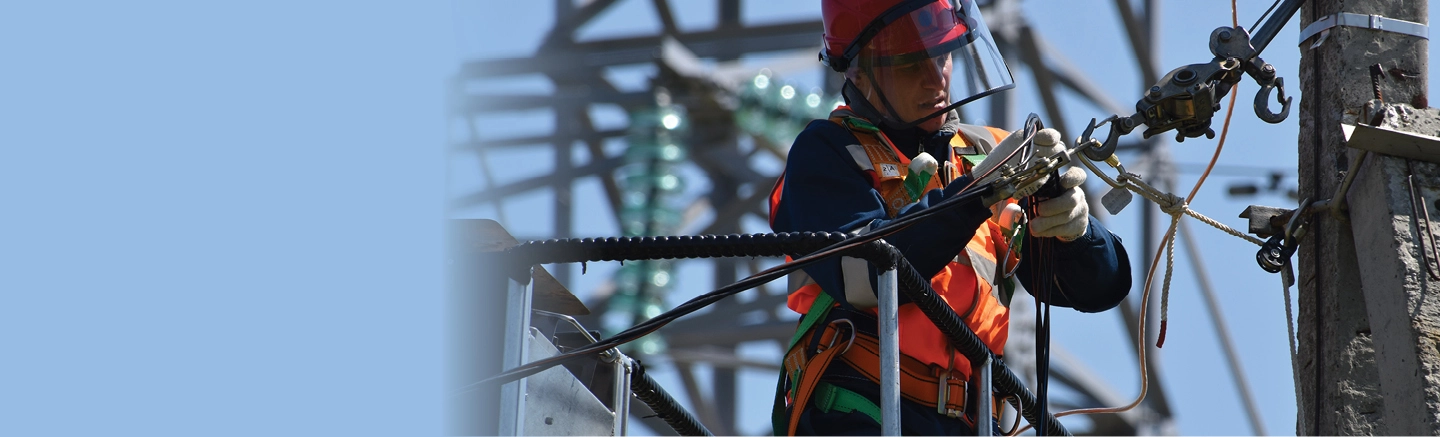 Utility worker wearing safety gear, including a red helmet and orange reflective vest, performing maintenance on electrical cables and equipment atop a utility pole. Metal structures and insulators are visible in the background.