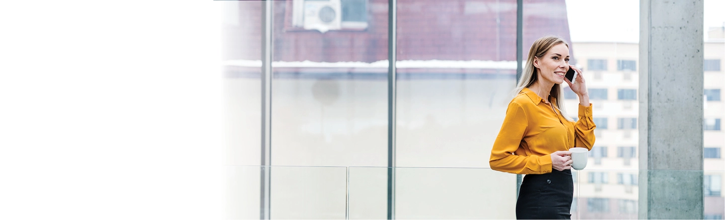 Professional in a modern office holding a coffee cup and talking on a mobile phone near large glass windows.