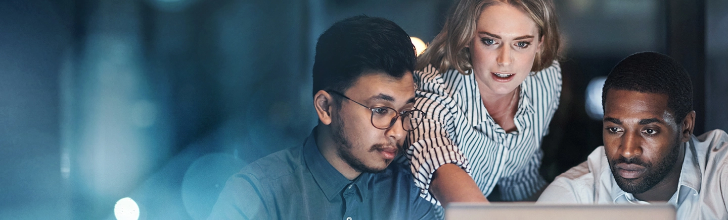 Team collaborating on a project using a laptop in a modern office environment with glass walls and evening lighting