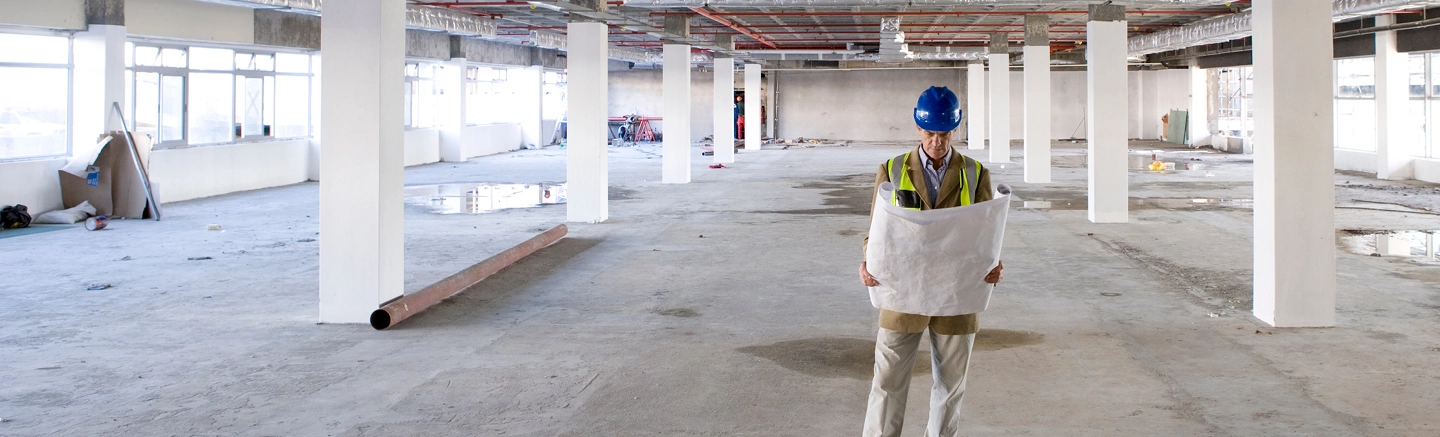 Construction worker wearing a blue safety helmet and reflective vest, standing in a large unfinished building interior while holding architectural plans. The space has concrete floors, white columns, and exposed ceiling pipes.