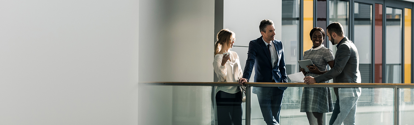 Four professionals standing and conversing in a modern office corridor with glass railings and large windows, holding documents and a tablet.