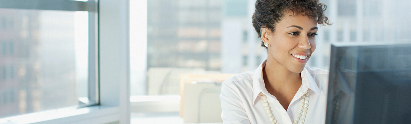 Person wearing a white shirt and pearl necklace working at a computer in a bright office with large windows and city buildings in the background.
