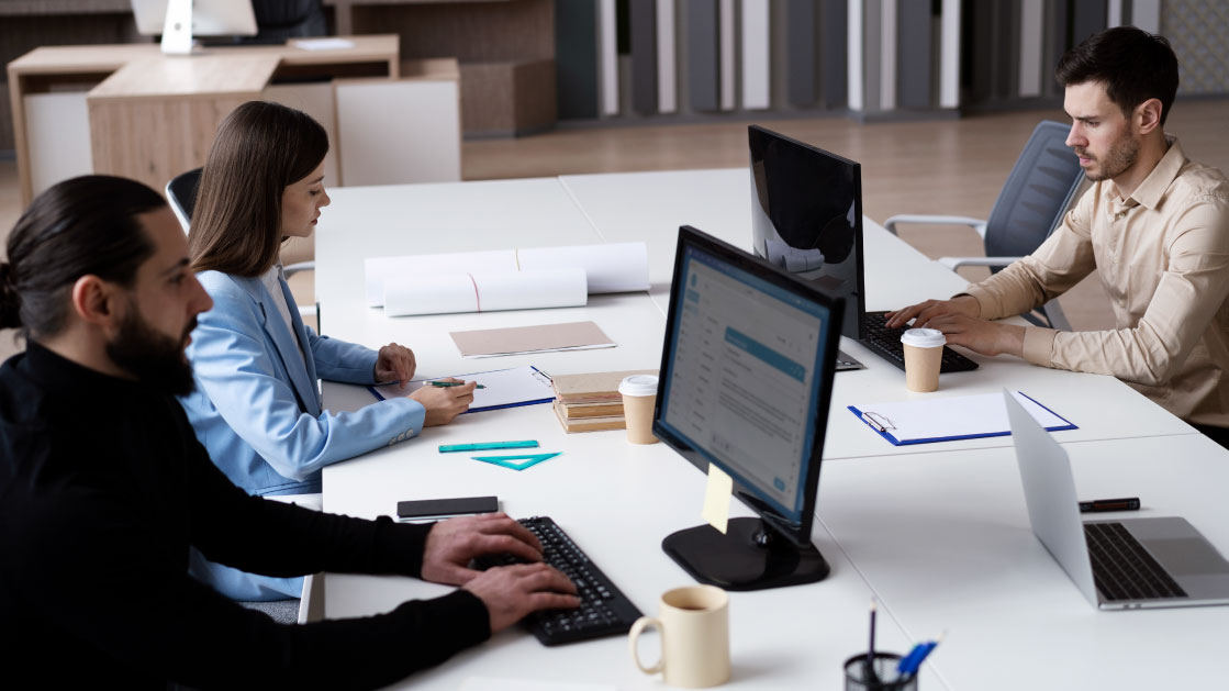 Un groupe de deux hommes et une femme travaillent sur un bureau. Les hommes travaillent sur des écrans tandis que la femme écrit sur un bloc-notes. Autour d'eux, il y a des tasses de café, un ordinateur portable, des papiers et un autre bloc-notes.