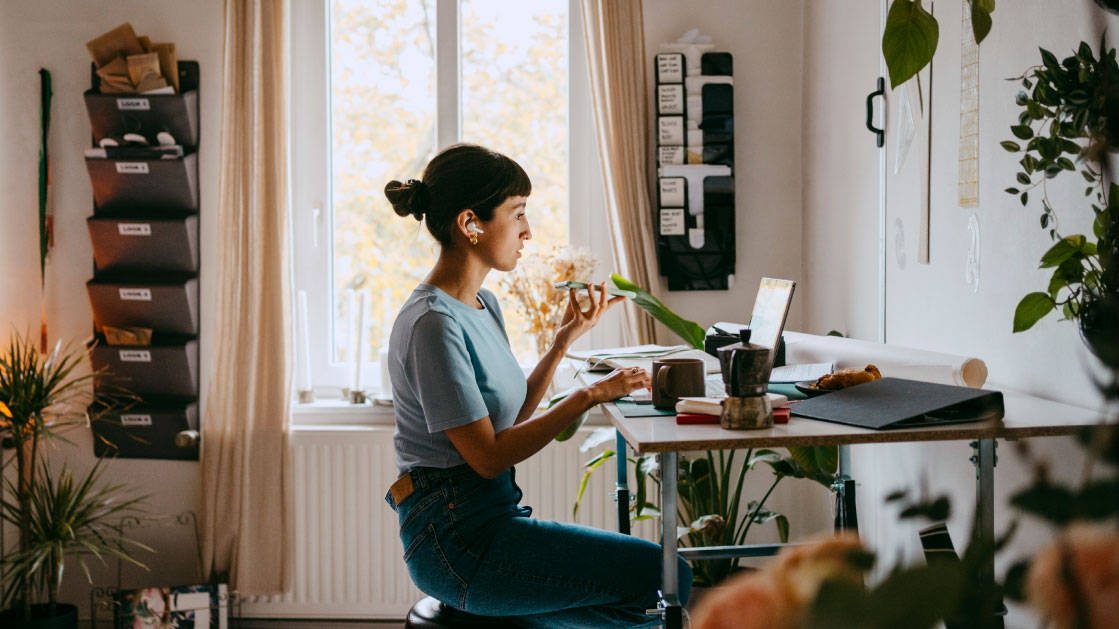 Une femme travaille chez elle, utilisant son ordinateur portable, ses écouteurs et parlant dans son téléphone portable. Elle se trouve dans un salon blanc entouré de plantes, avec un pot et une tasse de café.