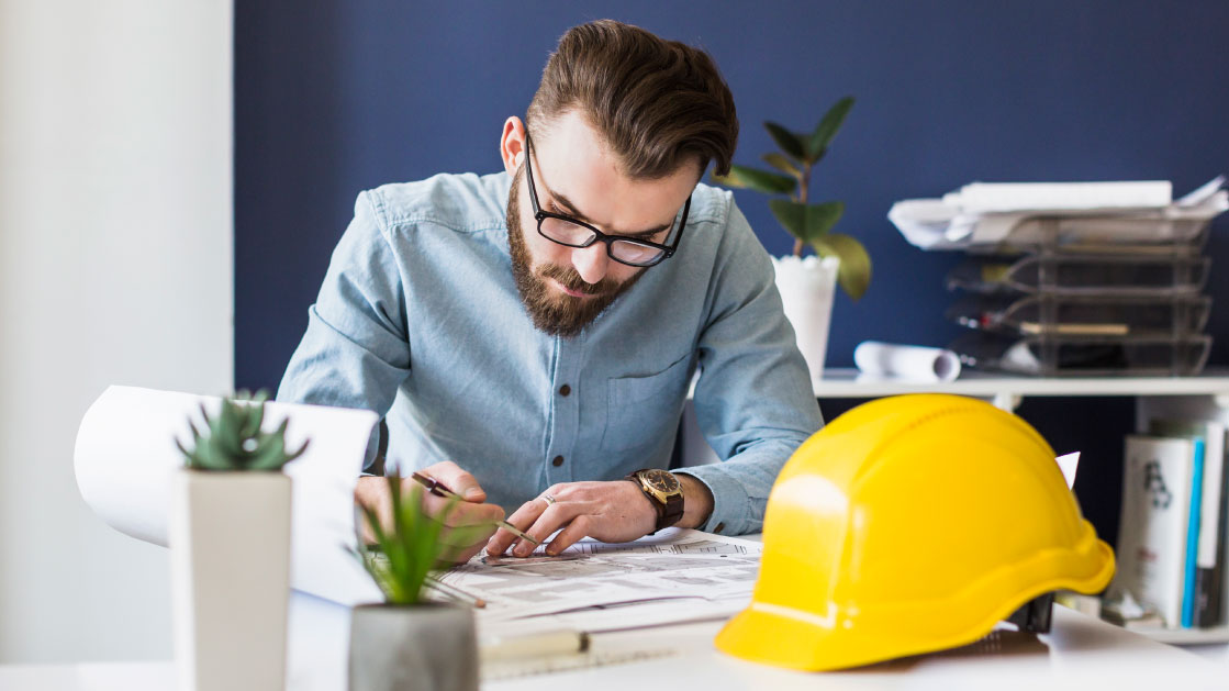 Un homme regarde des plans assis sur un bureau. Devant lui se trouve un casque de chantier jaune.