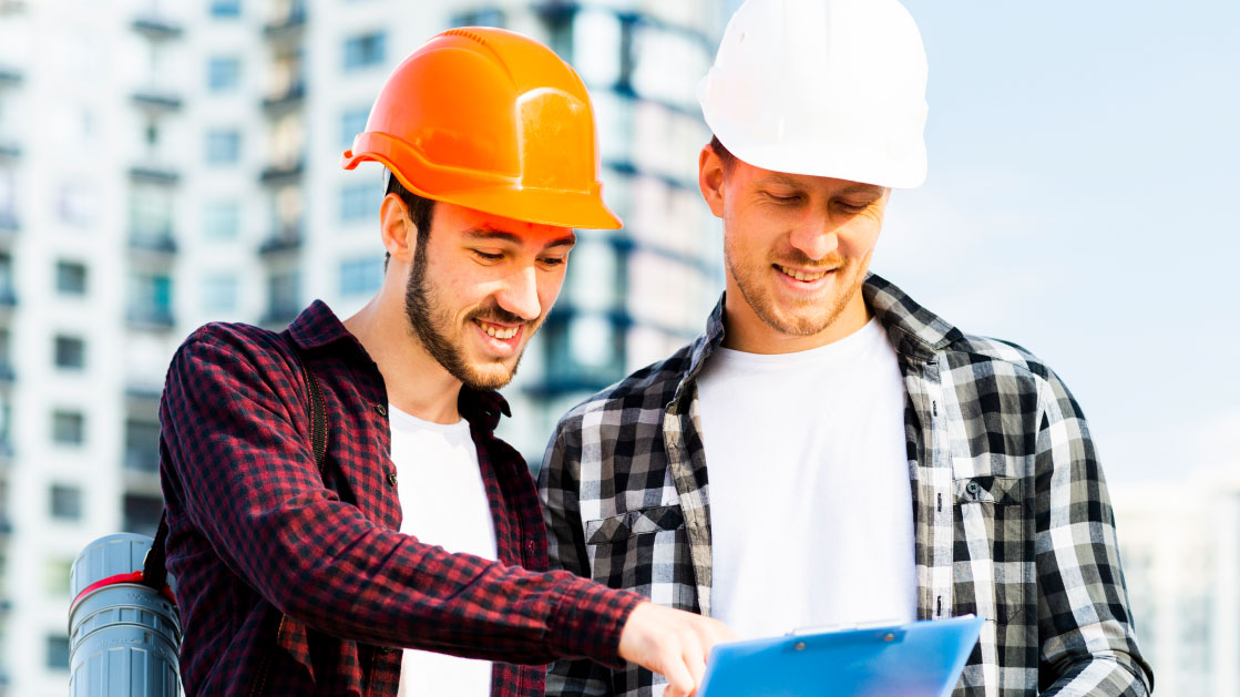 Deux hommes en chemises à carreaux et casques de chantier regardent un bloc-notes. Il y a un immeuble flou derrière eux.