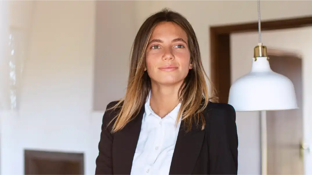 A woman in a white shirt and dark suit jacket smiles inside an office.