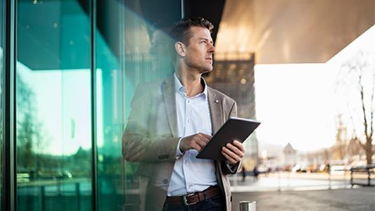 A man dressed in a blue shirt and tan suit jacket stands outside of an office while holding a tablet.