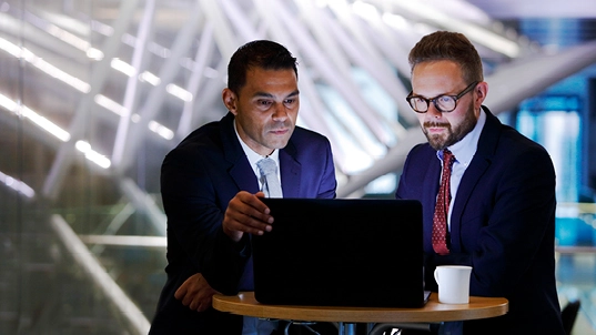 Deux hommes en costume bleu marine regardent un ordinateur. L'un d'eux porte une cravate rouge et des lunettes, l'autre une cravate gris-bleu.